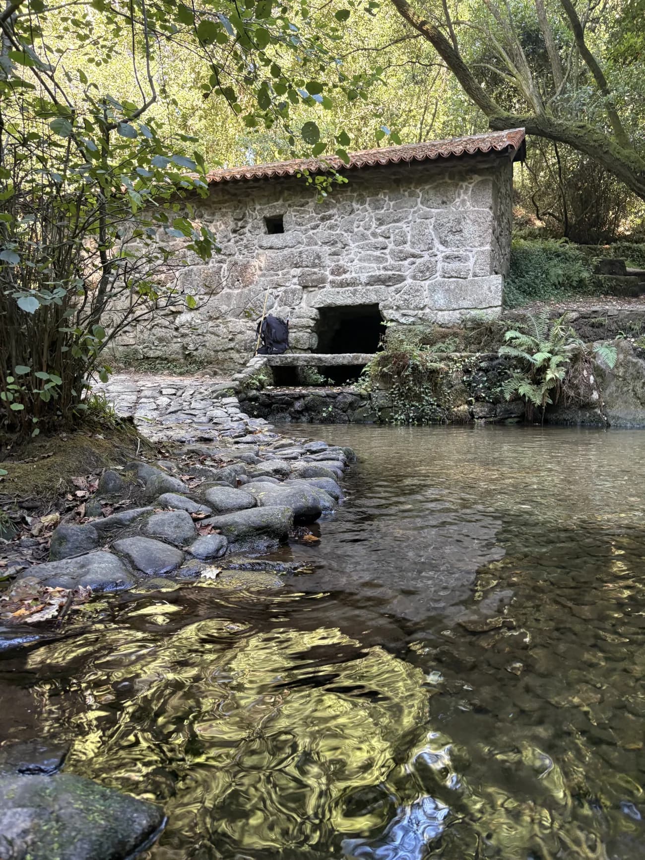 A stone watermill with a tiled roof beside a clear river, a pilgrim's pack and walking stick resting against its wall.