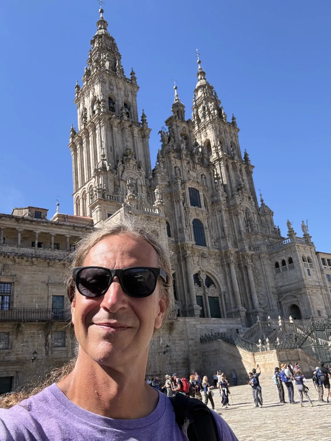 Mick Stemborski in sunglasses in front of the cathedral of Santiago de Compostela, pilgrims crossing the plaza behind him.