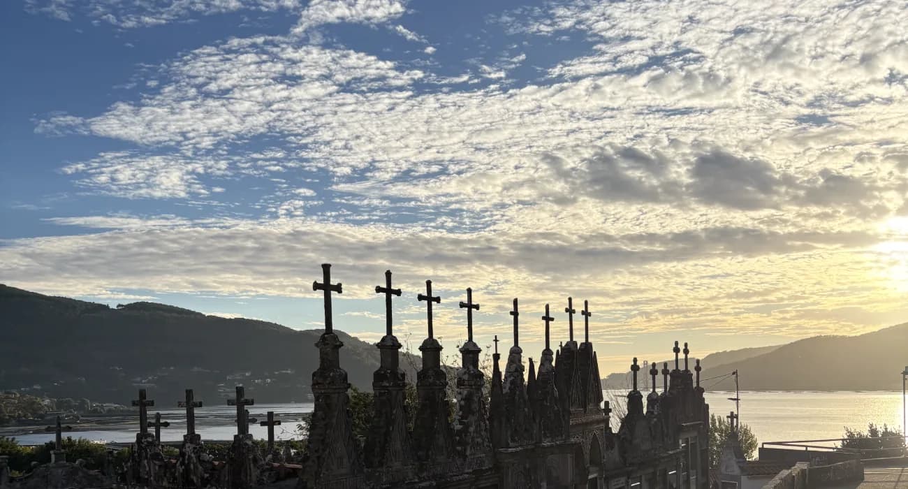 Stone crosses silhouetted at sunset along a hillside wall, water and hills beyond.