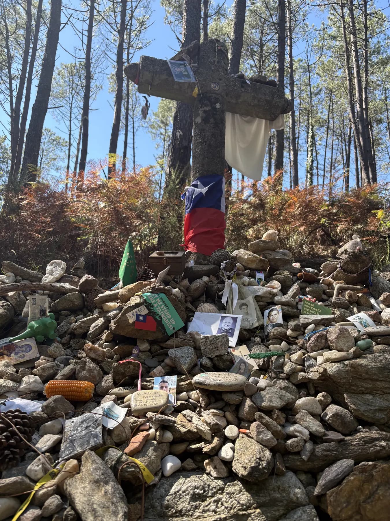 A wooden cross in a pine forest above a mound of stones left by pilgrims, with flags, photographs, and keepsakes tucked among them.